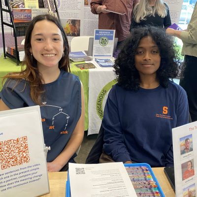 Two students sit behind a table at the Engaged Humanities Network Community Showcase, displaying linguistics teaching materials including a QR code poster and sentence diagrams. One wears a Mary Ann Shaw Center for Public and Community Service shirt.