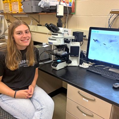 A student researcher (Emerson Long) poses next to a microscope and computer monitor displaying a microscopy image in a campus lab.