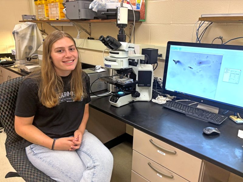 A student researcher (Emerson Long) poses next to a microscope and computer monitor displaying a microscopy image in a campus lab.