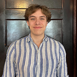 A young man with curly brown hair smiles in front of a dark wooden door, wearing a blue and white striped button-down shirt.
