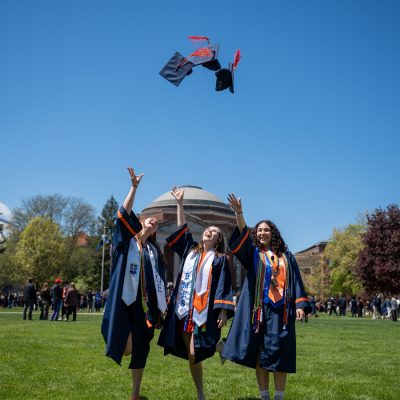 Three graduates in caps and gowns toss their mortarboards into the air on the Shaw Quad on a sunny day, with the Holden Observatory dome visible in the background.