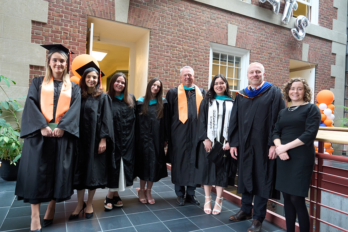 A group of eight people in graduation attire stand together in front of a brick building. Some are wearing mortarboards and stoles with colorful trims. There are orange and blue balloon decorations nearby, suggesting a festive graduation event