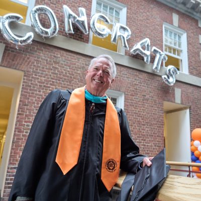 An individual in graduation attire stands smiling in front of a brick building. Above them, silver balloon letters spell CONGRATS.