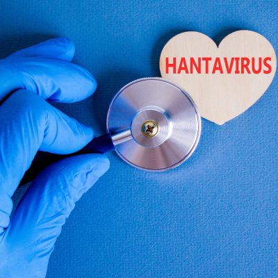 A gloved hand holds a stethoscope up to a wooden heart shape printed with the word 'Hantavirus' against a blue background