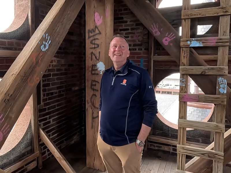 Person standing inside a brick tower structure with wooden beams and ladder, surrounded by circular window openings and colorful handprints painted on the wood.