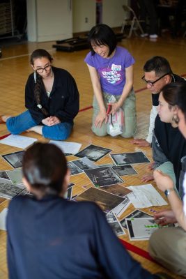 Four people sit on a gymnasium floor examining a spread of black-and-white historical photographs and documents, with one woman leaning in and gesturing as she leads the discussion.
