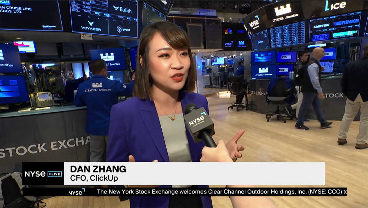 A person speaks into a NYSE microphone on the floor of the New York Stock Exchange, surrounded by trading screens and financial displays