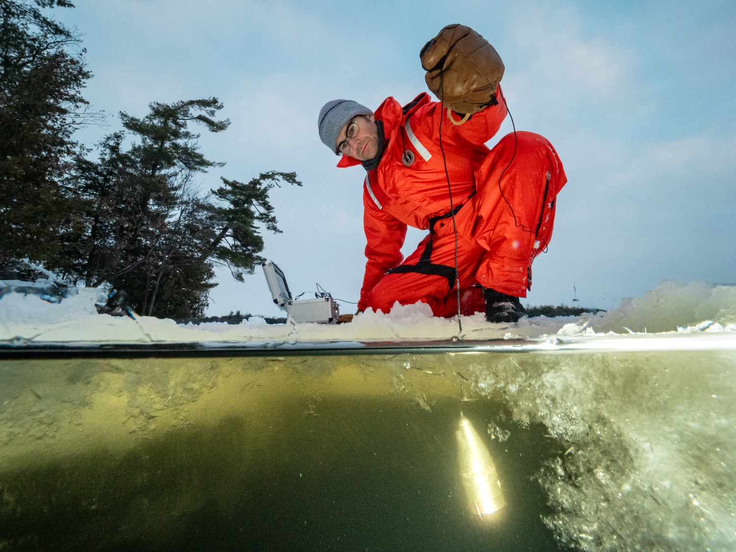 Person in red drysuit kneels on snowy lake shore, lowering a probe into an ice hole with half-above, half-below water view.