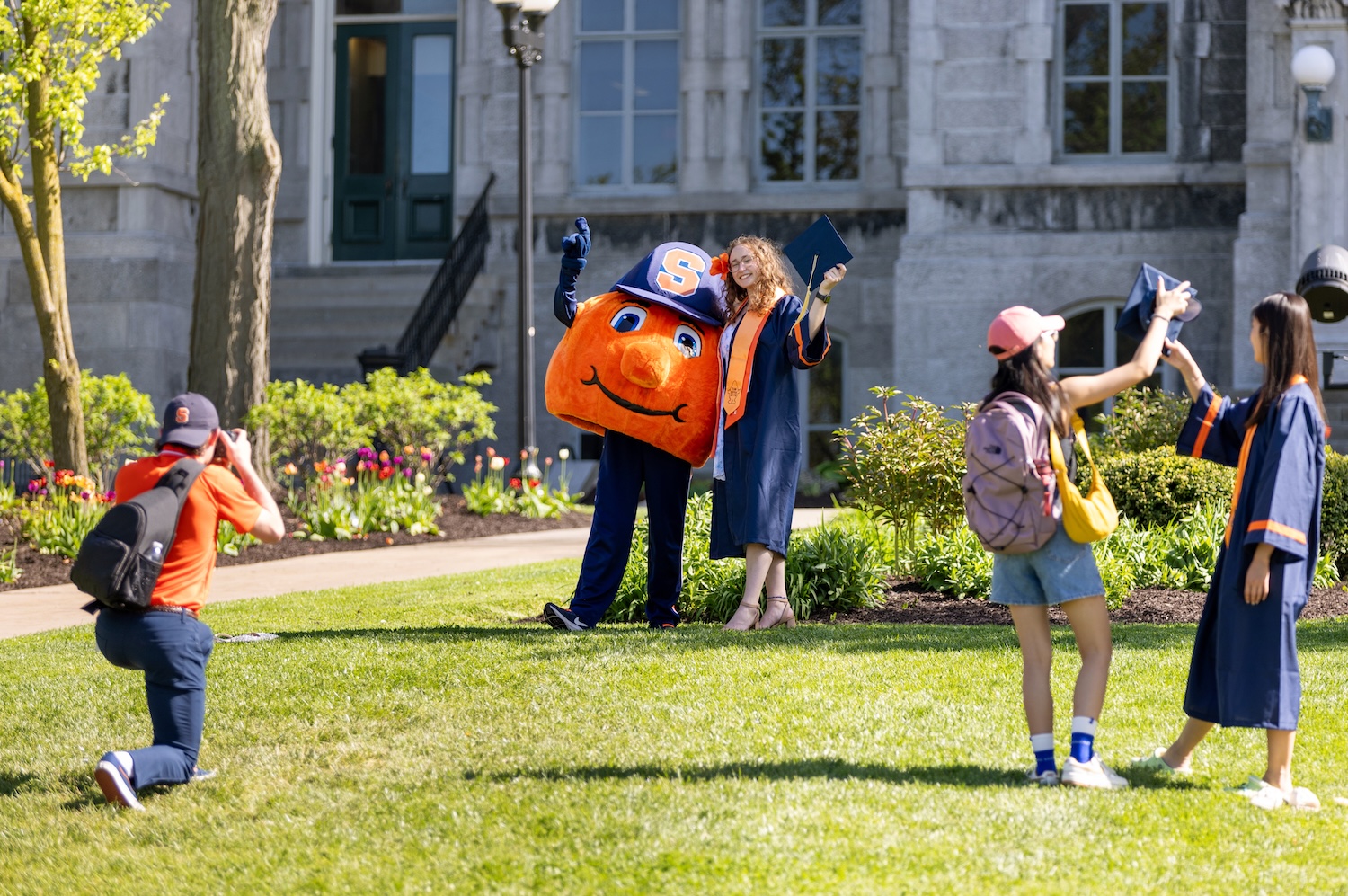 Graduate posing with an orange university mascot while others take photos on a sunny campus lawn outside a stone building.