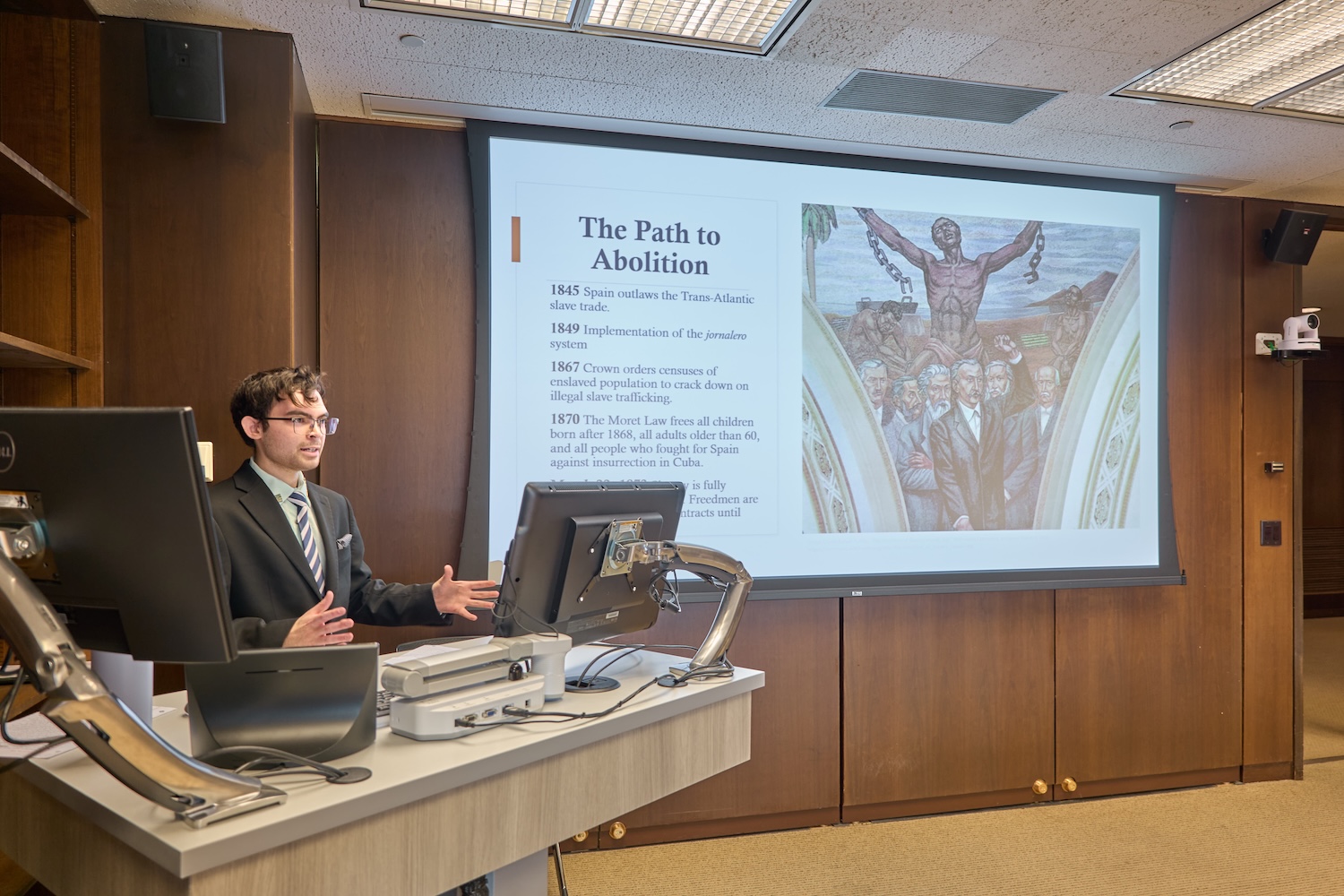 Person in a suit giving a classroom presentation at a lectern, with a slide titled “The Path to Abolition” projected on a screen.
