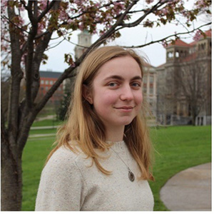 A young woman with long blonde hair smiles softly outdoors on a college campus, with a flowering tree and a historic brick building visible in the background.