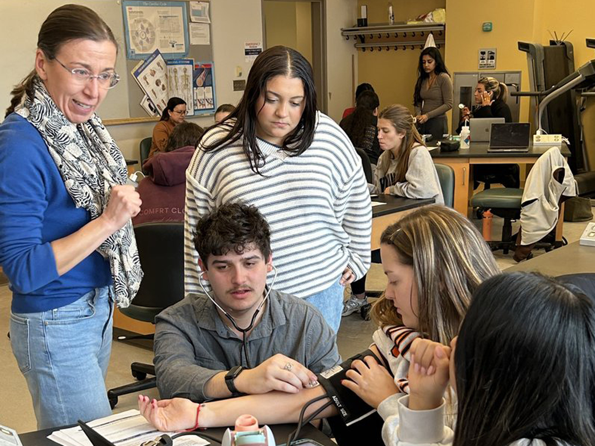 A professor instructs students during a lab exercise as a student uses a stethoscope to take a peer's blood pressure.