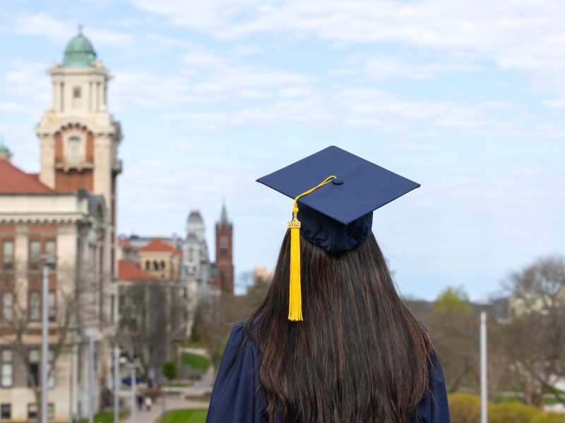 Back view of a graduate in cap and gown with a gold “2026” tassel, overlooking a campus with a domed building and cloudy sky.