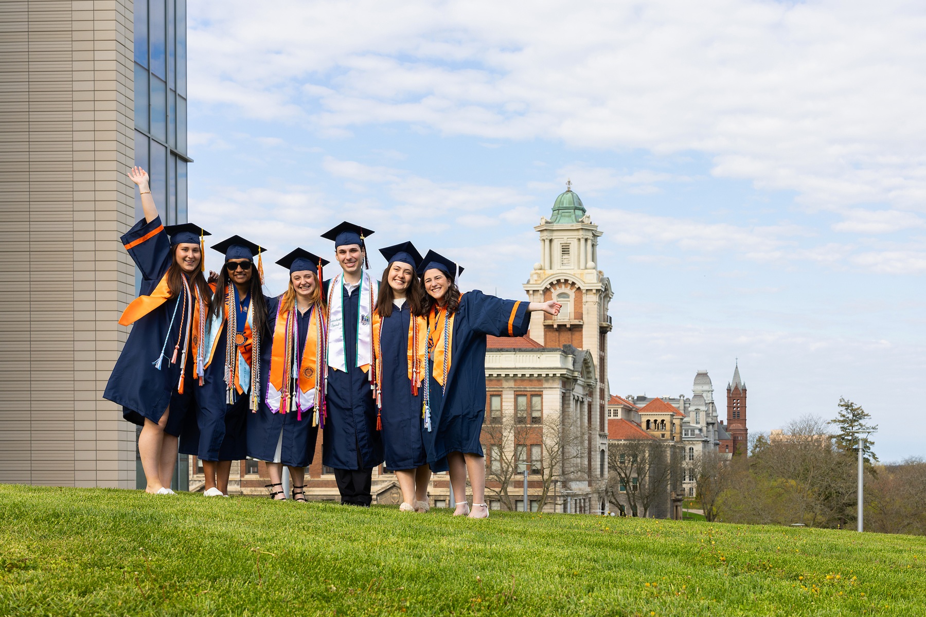 Six graduates in blue and orange regalia pose arm-in-arm on a green hillside 