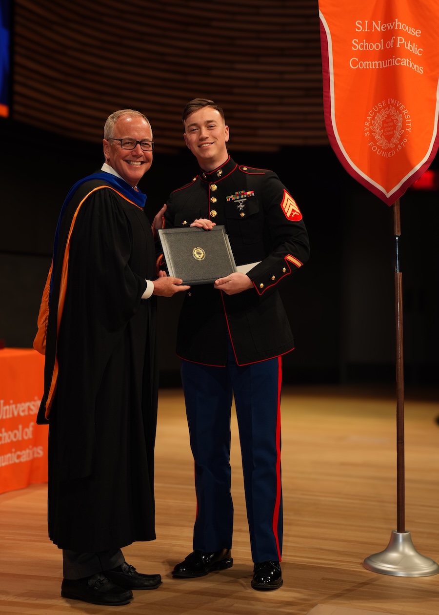 Graduate in military dress uniform receives a diploma onstage beside an academic official and a Syracuse University banner.