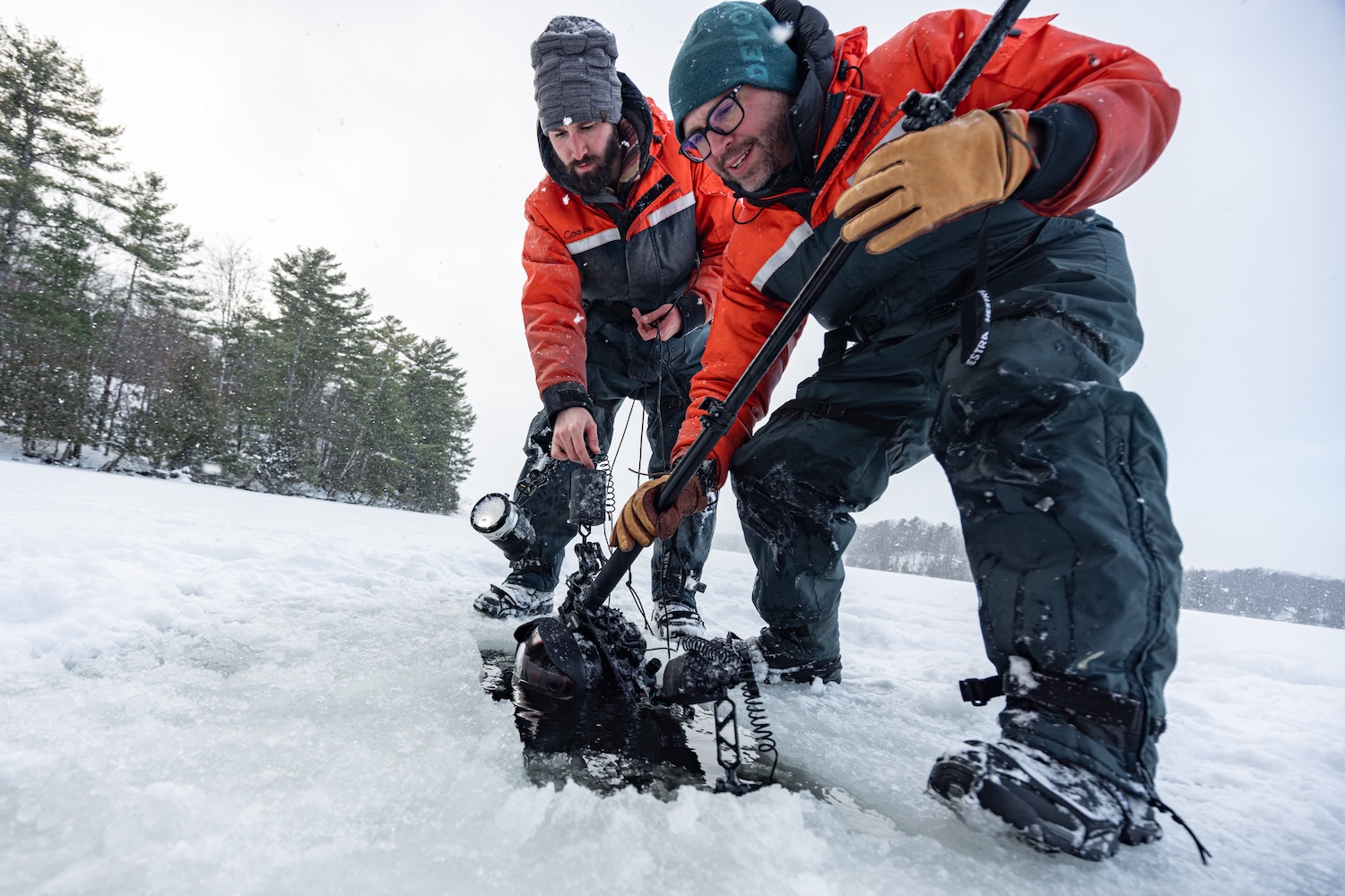 Two people in red drysuits kneel on a snowy frozen lake, lowering an underwater camera setup into a hole in the ice.