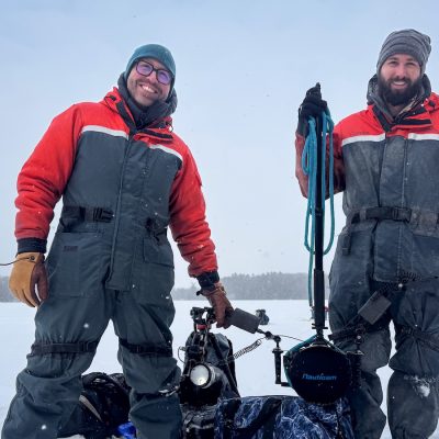 Two people in red drysuits stand on a snowy frozen lake beside underwater camera and lighting equipment on a sled during light snowfall.