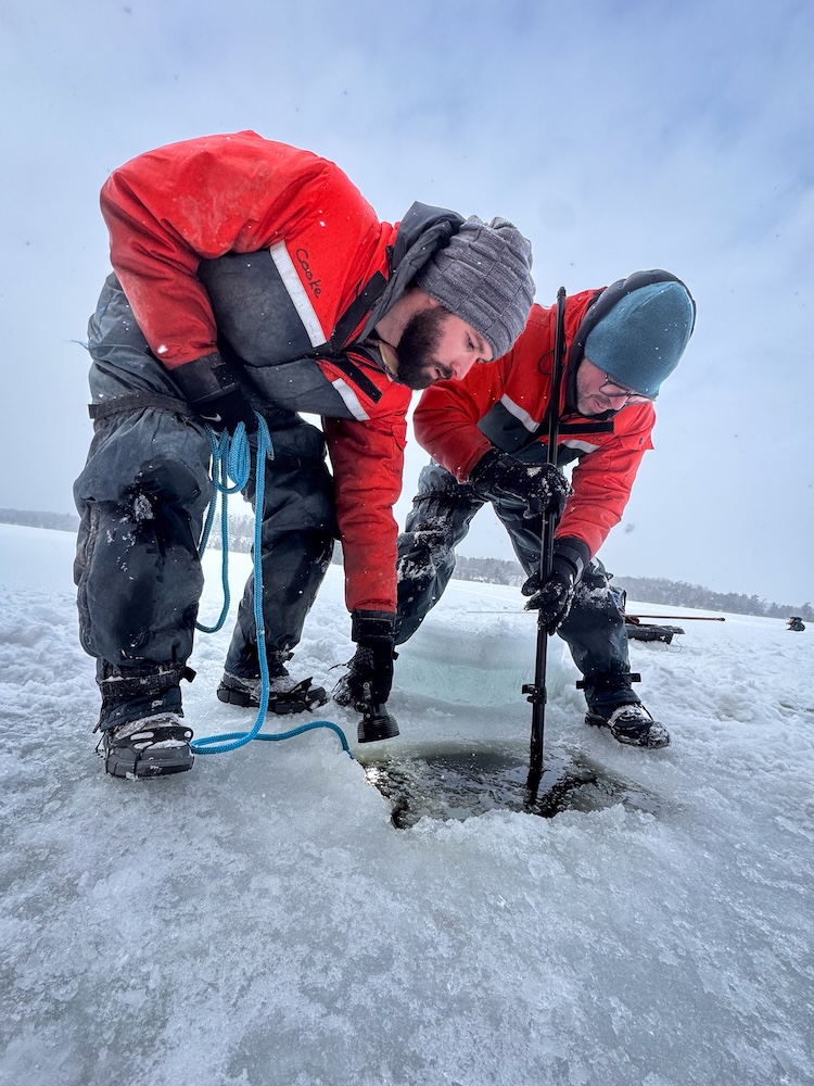 Two people in red drysuits kneel on a frozen lake, working with a probe and camera gear at a hole in the ice.