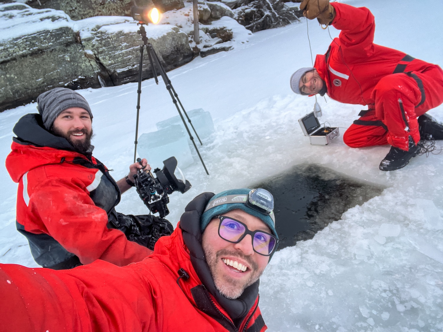 Three people in red suits work around an ice hole on a frozen lake, with camera equipment and a tripod nearby.