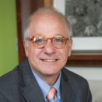 A man in a gray blazer, blue shirt and orange tie smiles in a portrait setting.