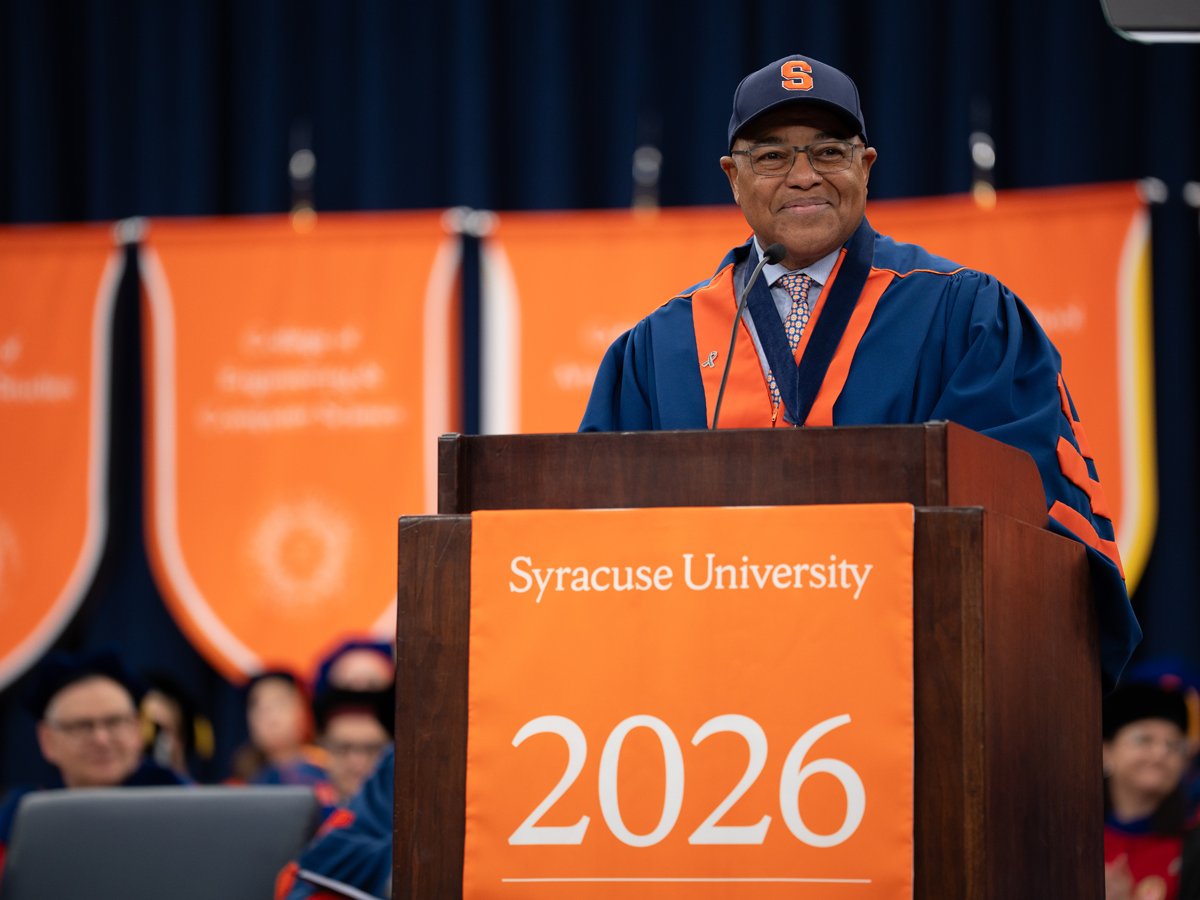 The Commencement speaker delivers remarks at the podium wearing a navy Syracuse University cap.