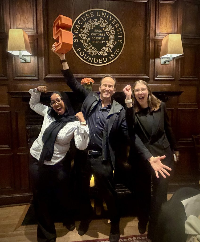 Three people celebrate exuberantly in front of a Syracuse University seal on a wood-paneled wall, with one person raising an orange block-letter 'S' trophy above their head