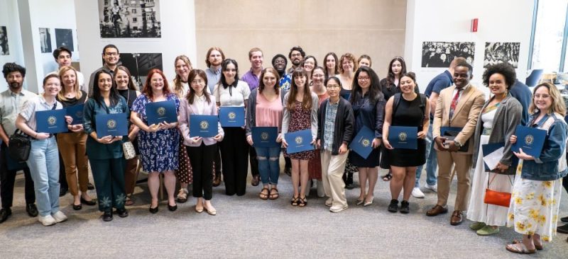A group of approximately 25 people smile for a photo, several holding navy blue certificate folders, at an academic awards ceremony.