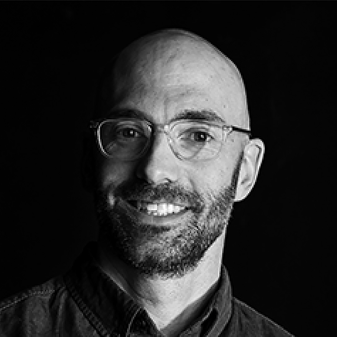 Black-and-white headshot of a person with glasses and a beard against a dark background.