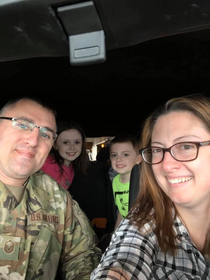 Four people smiling inside a car; one person in a U.S. Air Force uniform, two children in the back seat, and another adult in the front.
