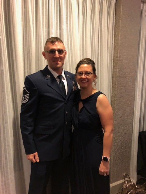 Two people standing side by side indoors in formal attire, one wearing a U.S. Air Force dress uniform, in front of light-colored curtains.