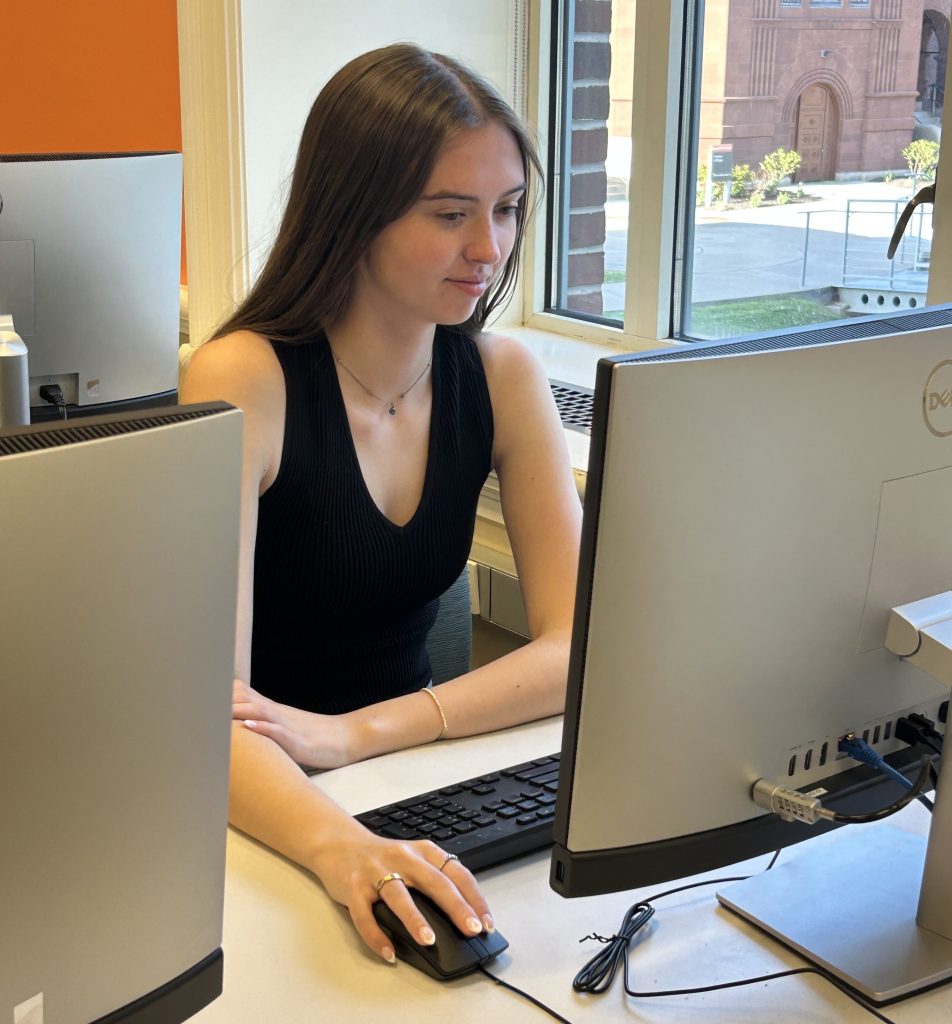 A young woman with long brown hair works at a desktop computer in a campus computer lab, with a red brick building visible through the window behind her.