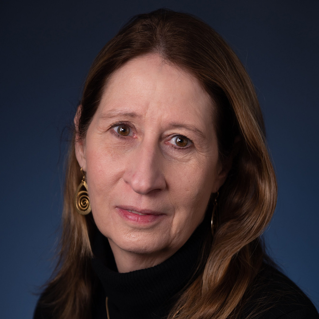 Studio headshot of a person with long hair and dangling earrings against a blue background.