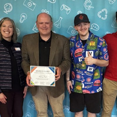 A faculty member holds a SOURCE Award certificate while posing with three others in front of a teal activity-themed backdrop.