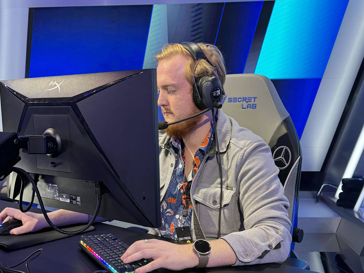 A student wears a headset and works at a gaming station, with an esports arena backdrop behind him.
