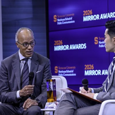Two people in suits sit onstage holding microphones during an interview, with a “2026 Mirror Awards” Syracuse University backdrop behind them.
