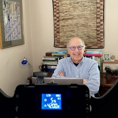 A man smiles with arms crossed at a desk, seen through a ring light and camera setup in a home office.