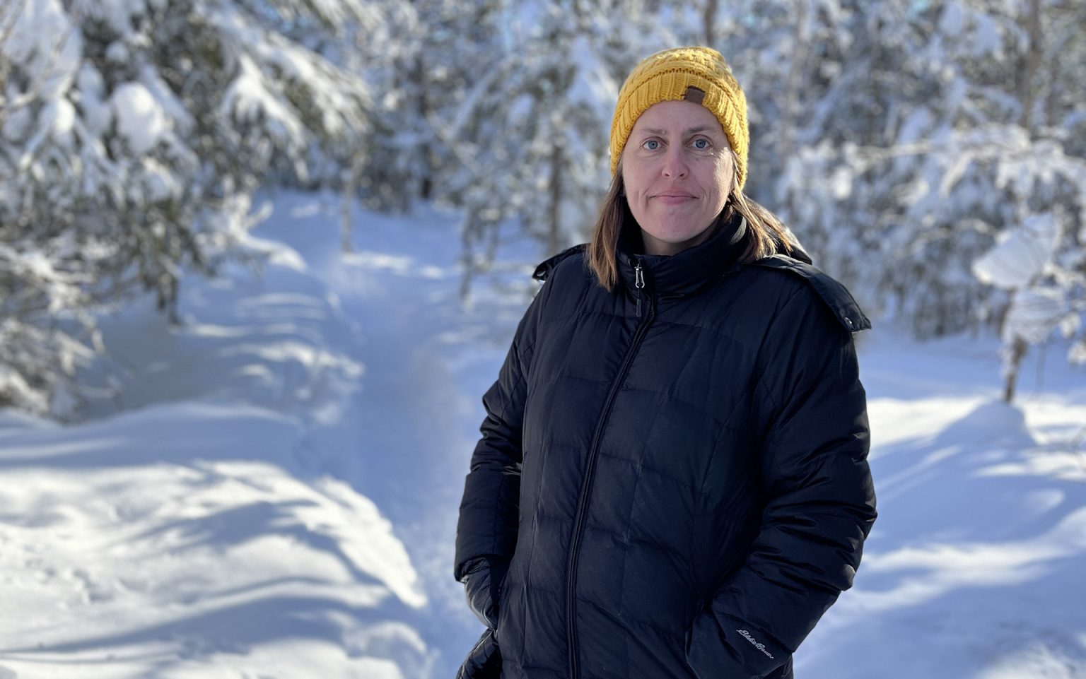 A person stands on a snow-covered trail in a black jacket and yellow knit hat.