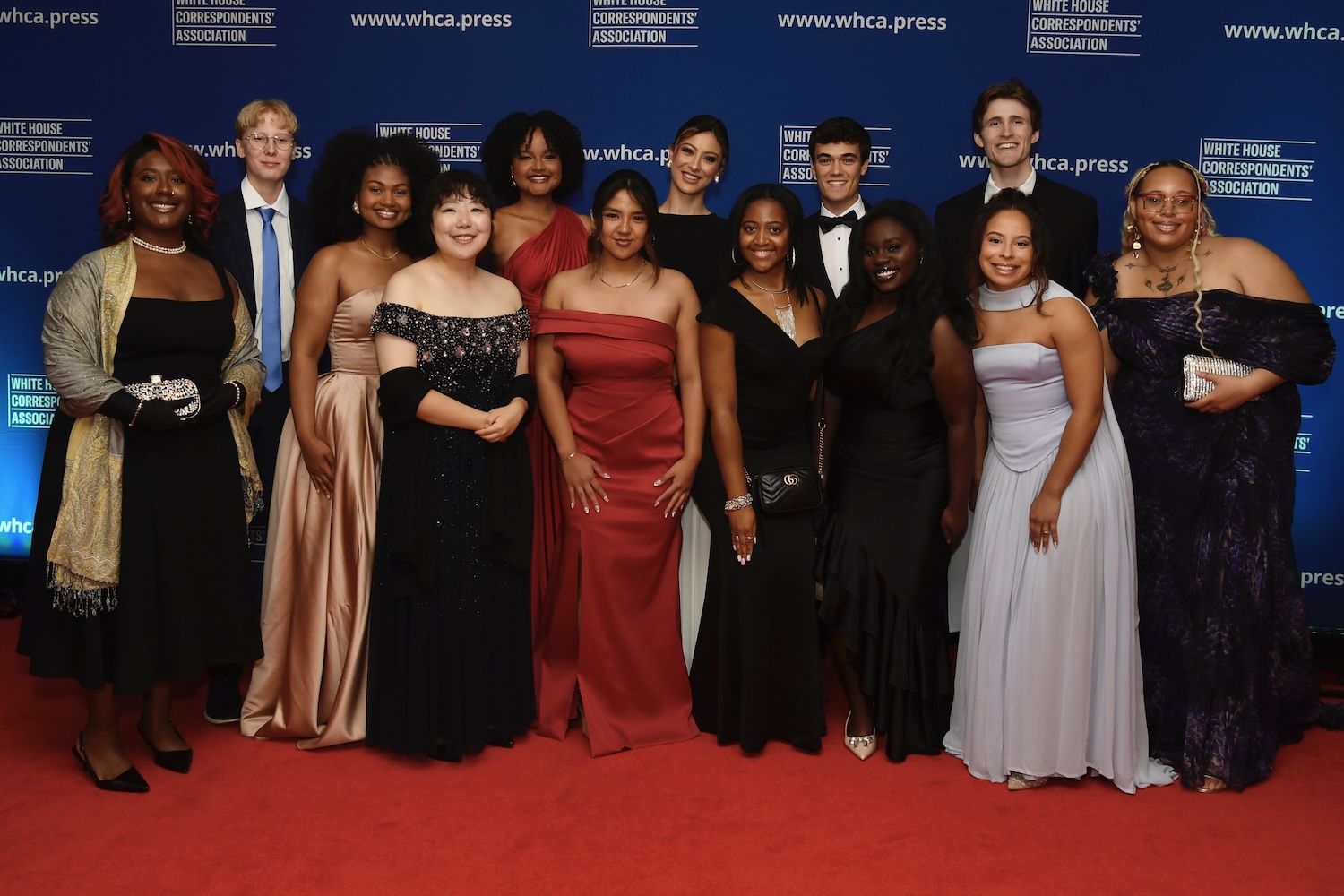 Group of formally dressed attendees posing on a red carpet in front of a blue “White House Correspondents’ Association” backdrop.