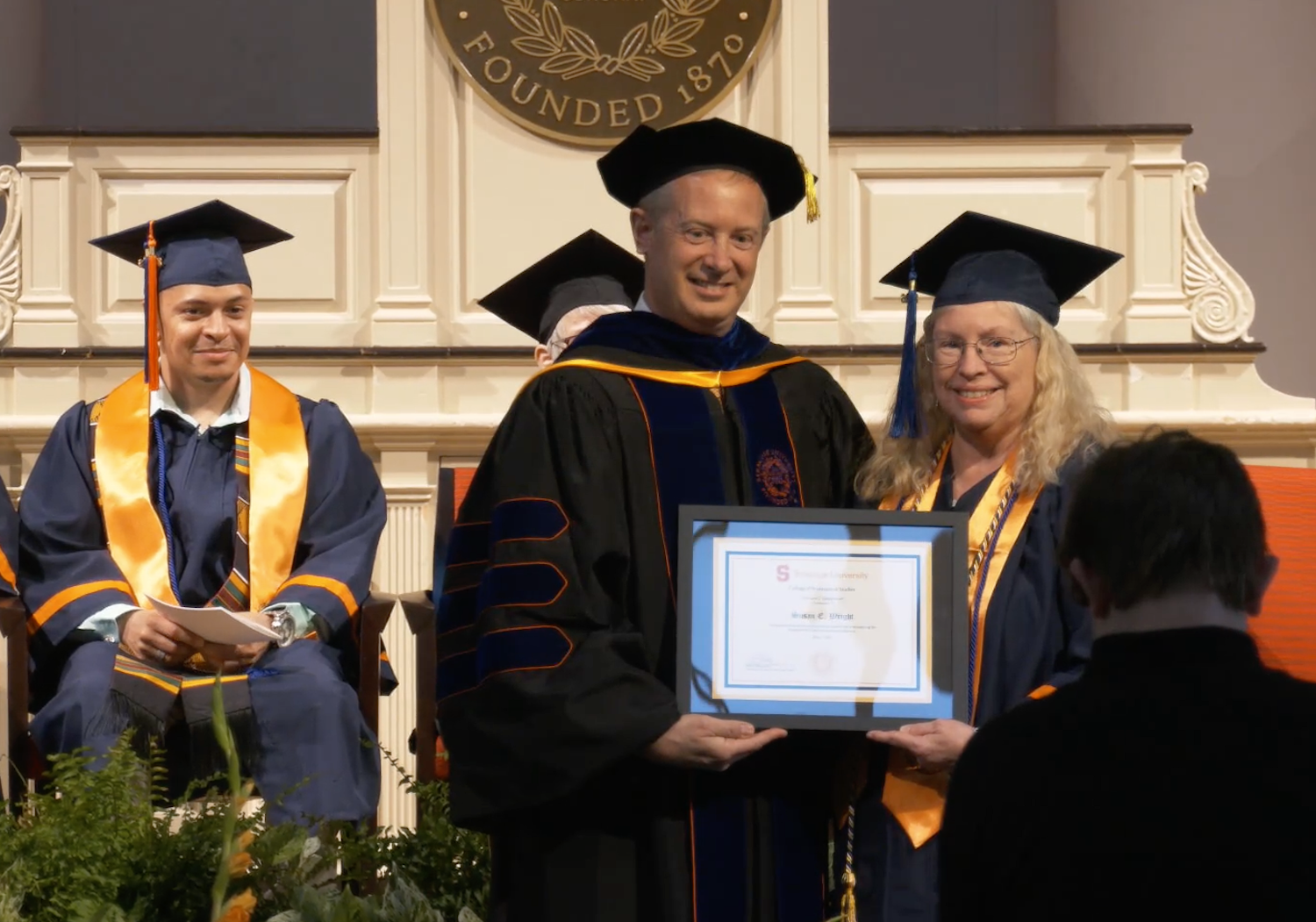Graduate in cap and gown receives framed Syracuse University award on stage during commencement ceremony.