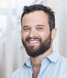 A man with short dark hair and a beard smiles at the camera, wearing a light blue shirt against a softly blurred background.