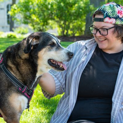 A person smiles while sitting on the grass next to a large dog wearing a pink and black harness on a sunny day on the Syracuse University campus, with green lawns and stone buildings in the background.