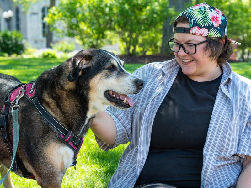 A person smiles while sitting on the grass next to a large dog wearing a pink and black harness on a sunny day on the Syracuse University campus, with green lawns and stone buildings in the background.