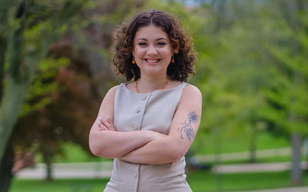 A person smiles with arms crossed, wearing a gray sleeveless outfit outdoors.