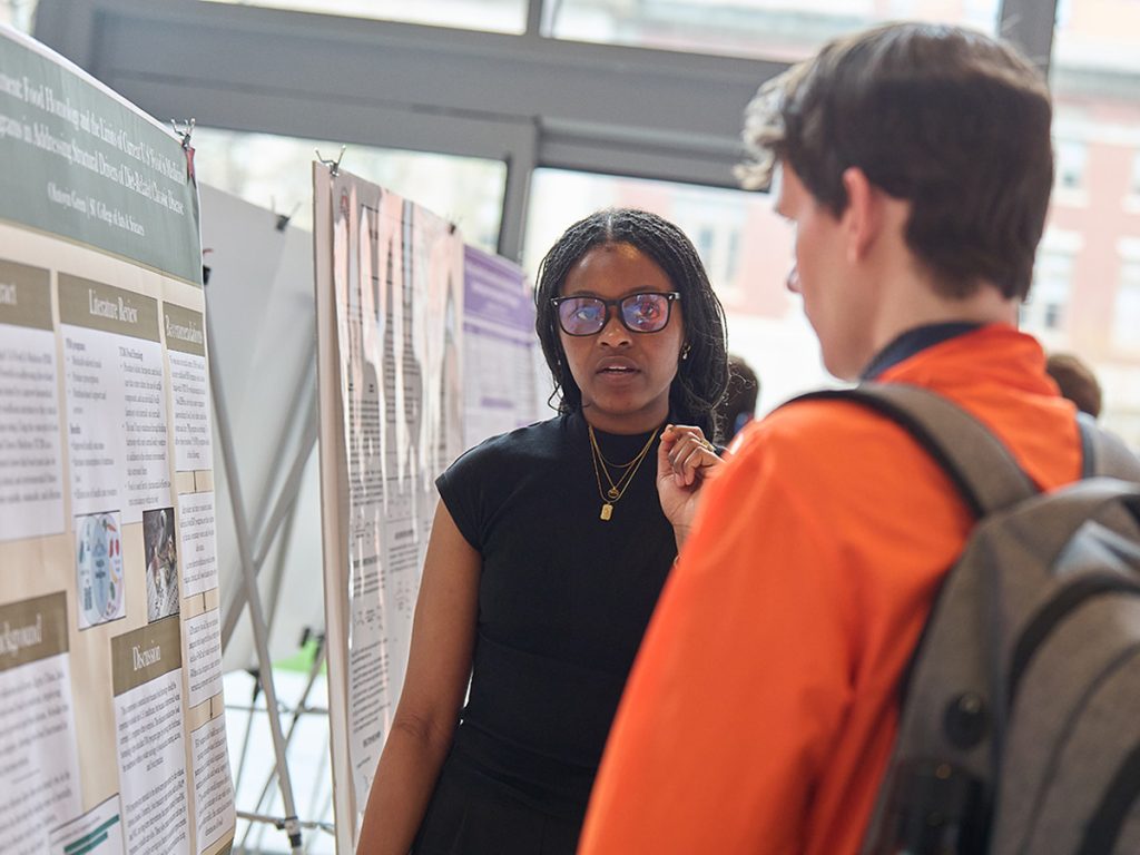 A student wearing glasses presents her research poster to an attendee at a university research festival, gesturing as she explains her work on food insecurity and diet-related chronic disease.