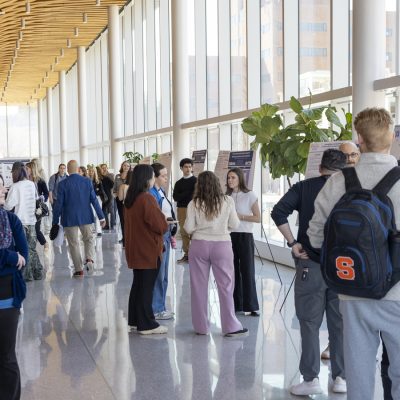 Students and faculty browse research posters in a sunlit atrium during an academic poster session.