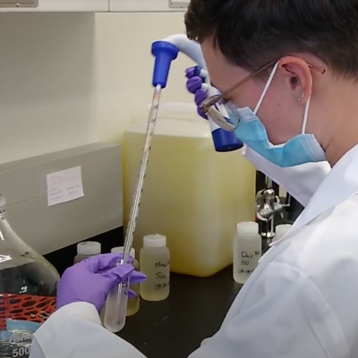 Person in a lab coat, gloves, and mask uses a pipette to transfer liquid into a test tube at a laboratory bench with bottles and a large flask.