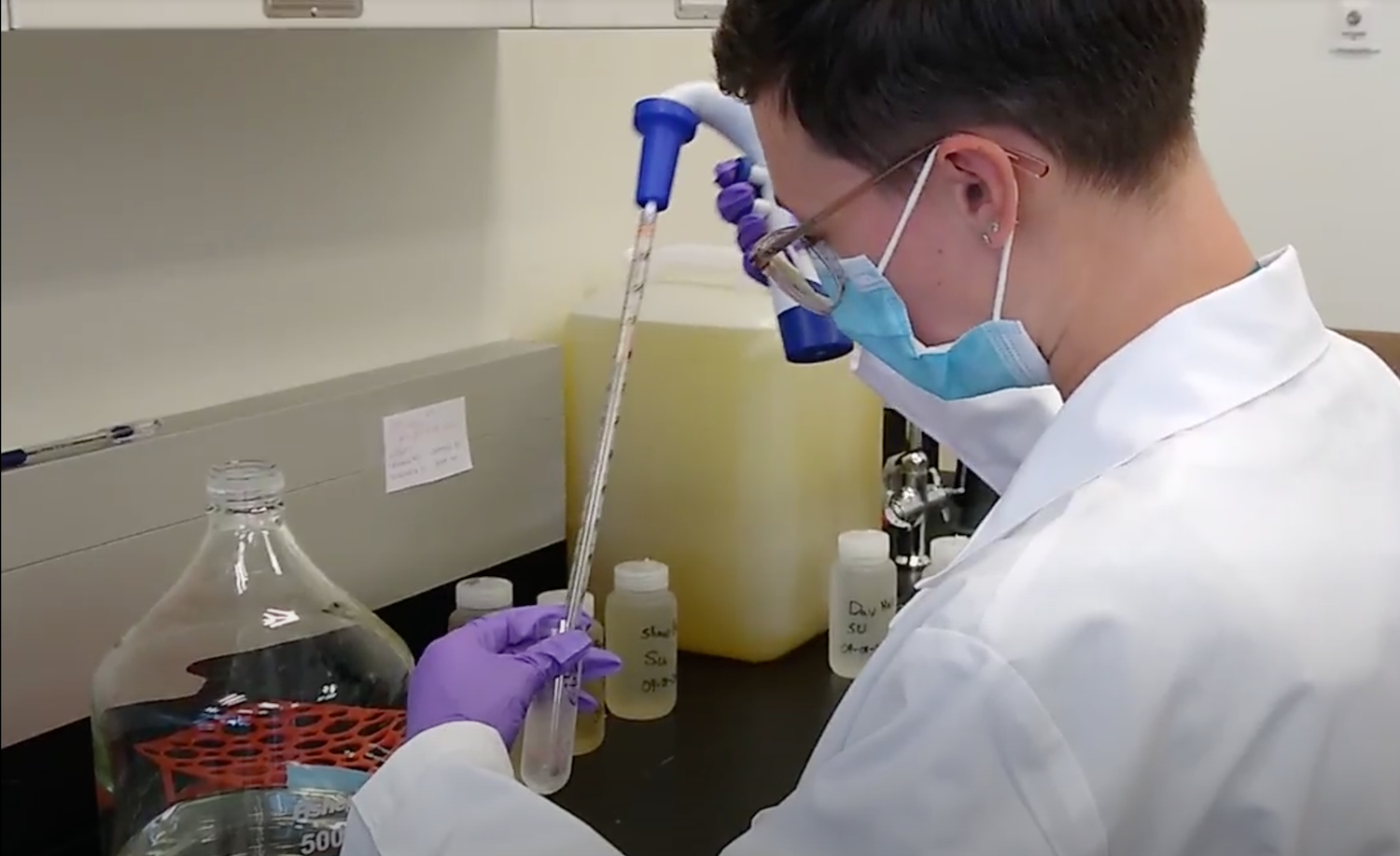 Person in a lab coat, gloves, and mask uses a pipette to transfer liquid into a test tube at a laboratory bench with bottles and a large flask.