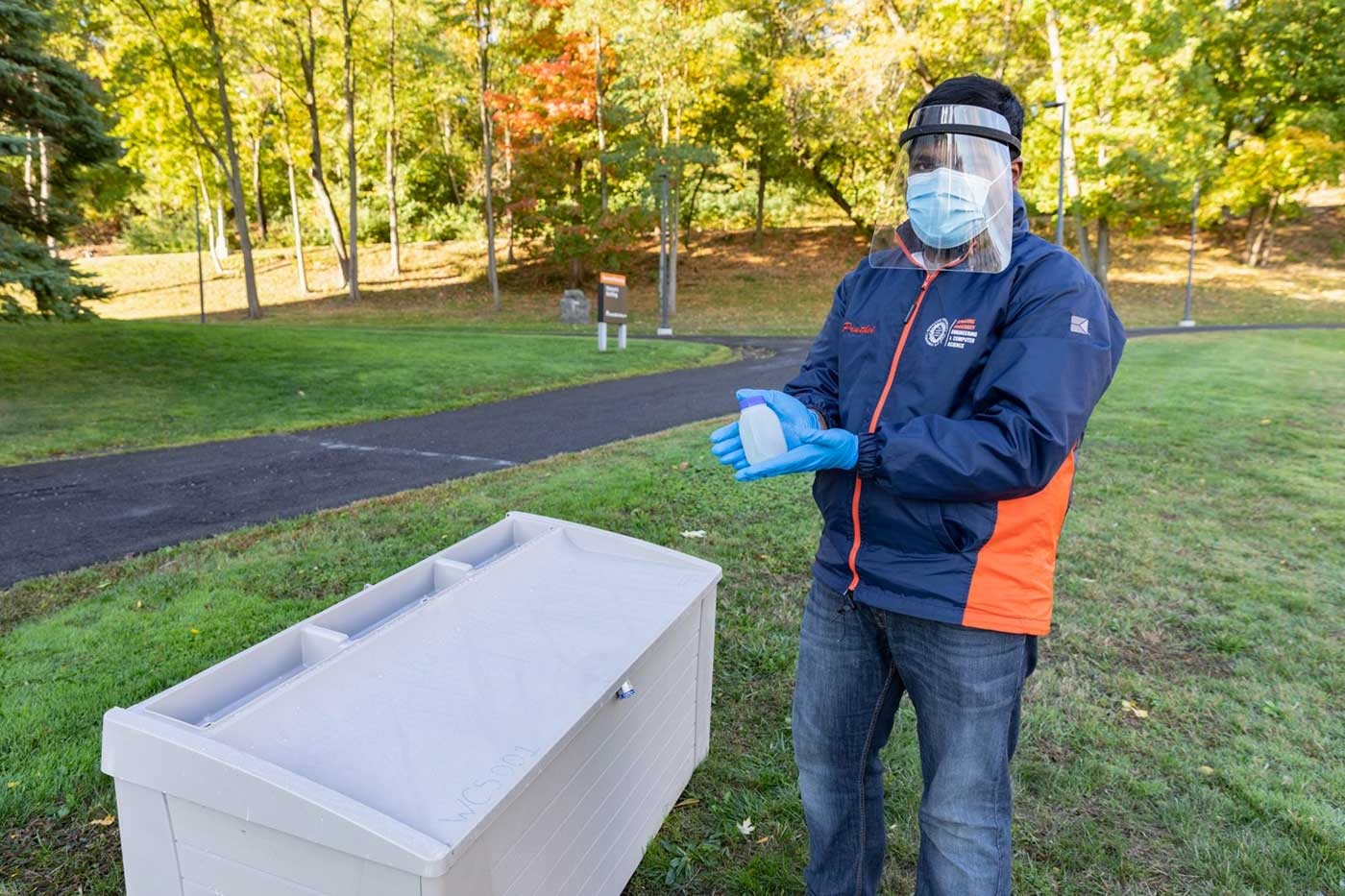 Person wearing a face shield, mask, and gloves holds a sample container beside a gray collection bin in an outdoor setting.
