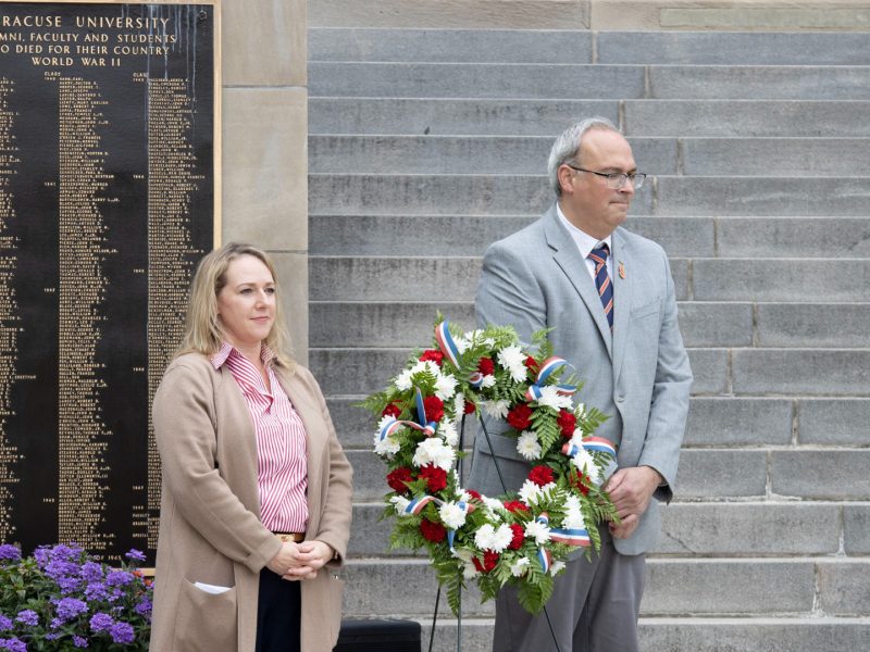 Two people stand in front of the Syracuse University World War II memorial plaque on the steps of Hendricks Chapel during a wreath-laying ceremony. A man holds a red, white and blue floral wreath while a woman stands beside him with hands clasped. The bronze plaque behind them lists the names of alumni, faculty and students who died in World War II.