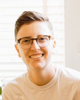 A person with short hair and tortoiseshell glasses smiles warmly at the camera in a bright, naturally lit setting.